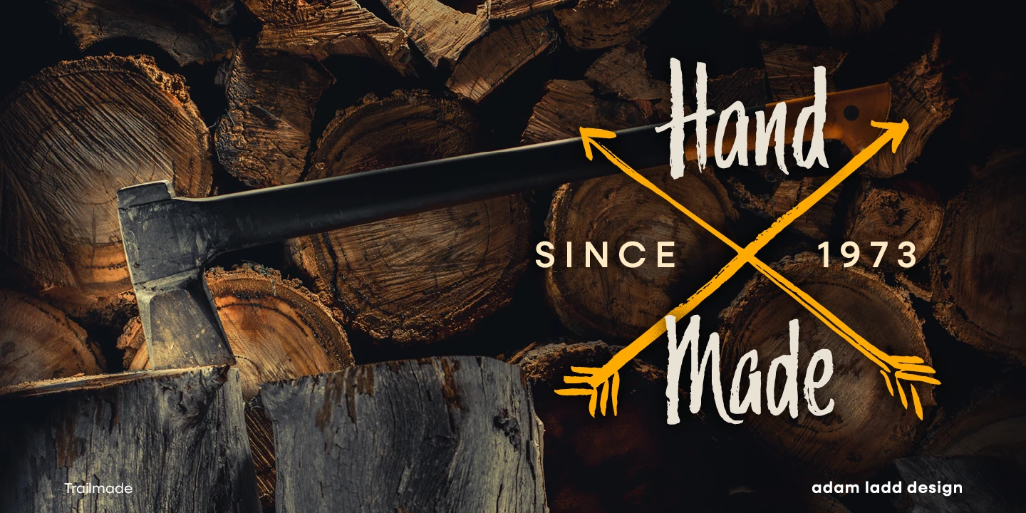 Trailmade font by Adam Ladd Design — the words “Hand Made” in a connected script, crossed by two yellow arrows, with “SINCE” and “1973” in sans-serif capitals, all over a pile of logs with an axe stuck in one